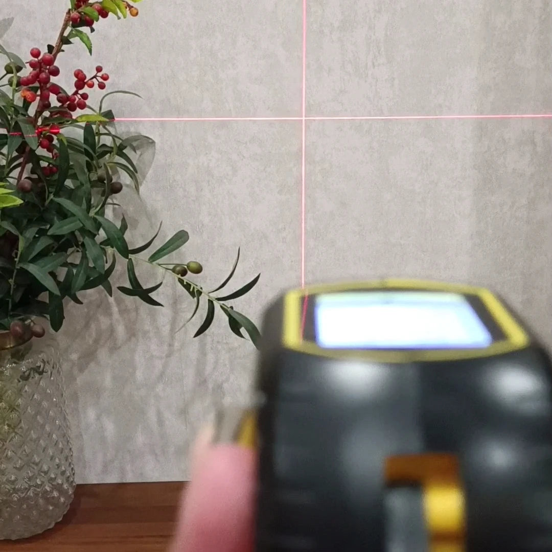 Digital 3-in-1 laser distance meter with laser crosshair targeting a grey wall near a vase with green leaves and berries