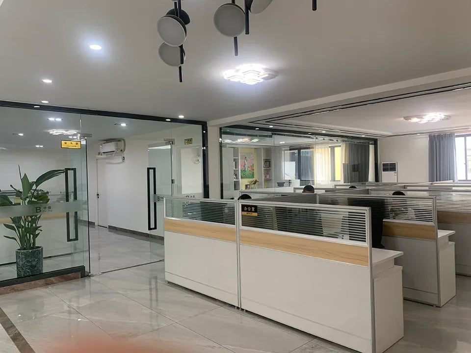 Modern office space with glass walls, white desks, partition panels, and ceiling light fixtures in a bright room