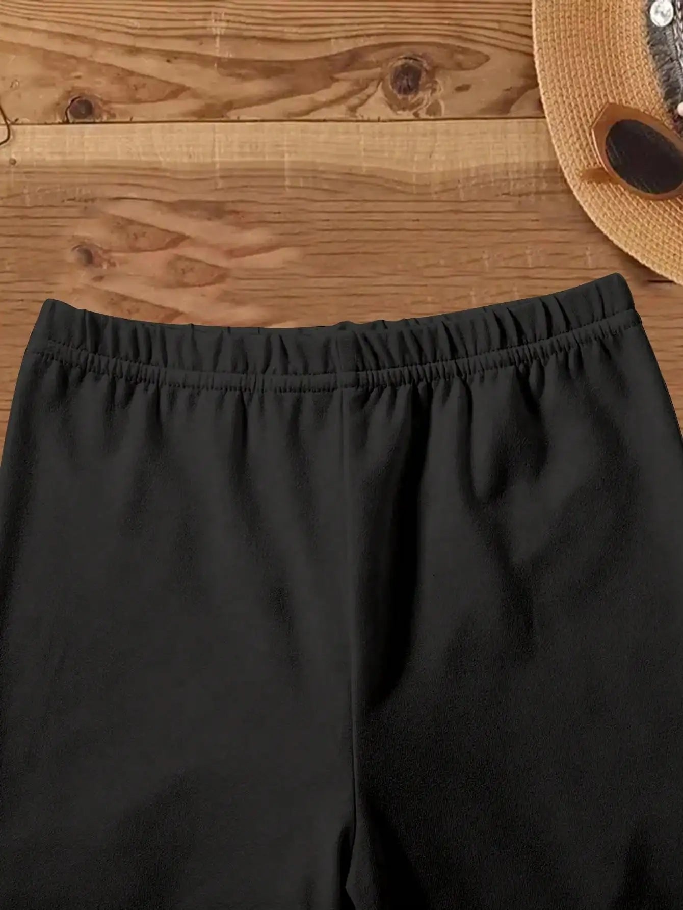 Close-up of black skinny shorts with elastic waistband displayed on wooden surface with hat and sunglasses nearby