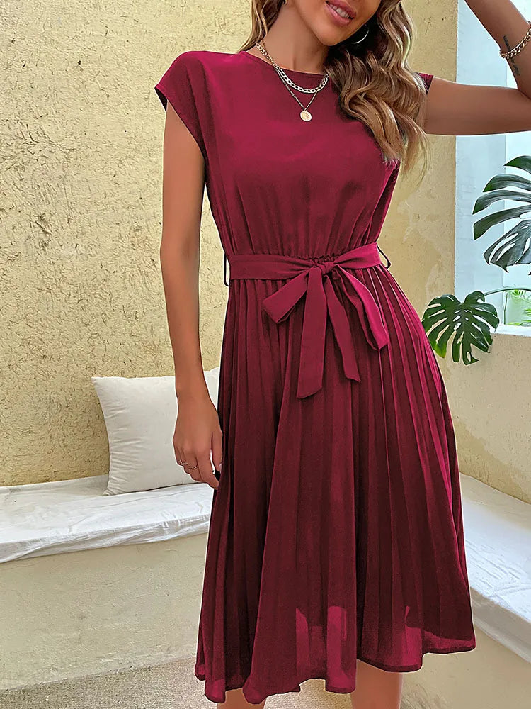 Woman wearing elegant sleeveless burgundy pleated summer dress with waist tie in bright indoor setting