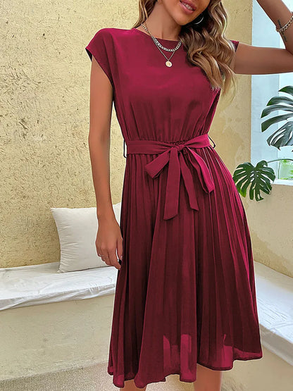 Woman wearing elegant sleeveless burgundy pleated summer dress with waist tie in bright indoor setting