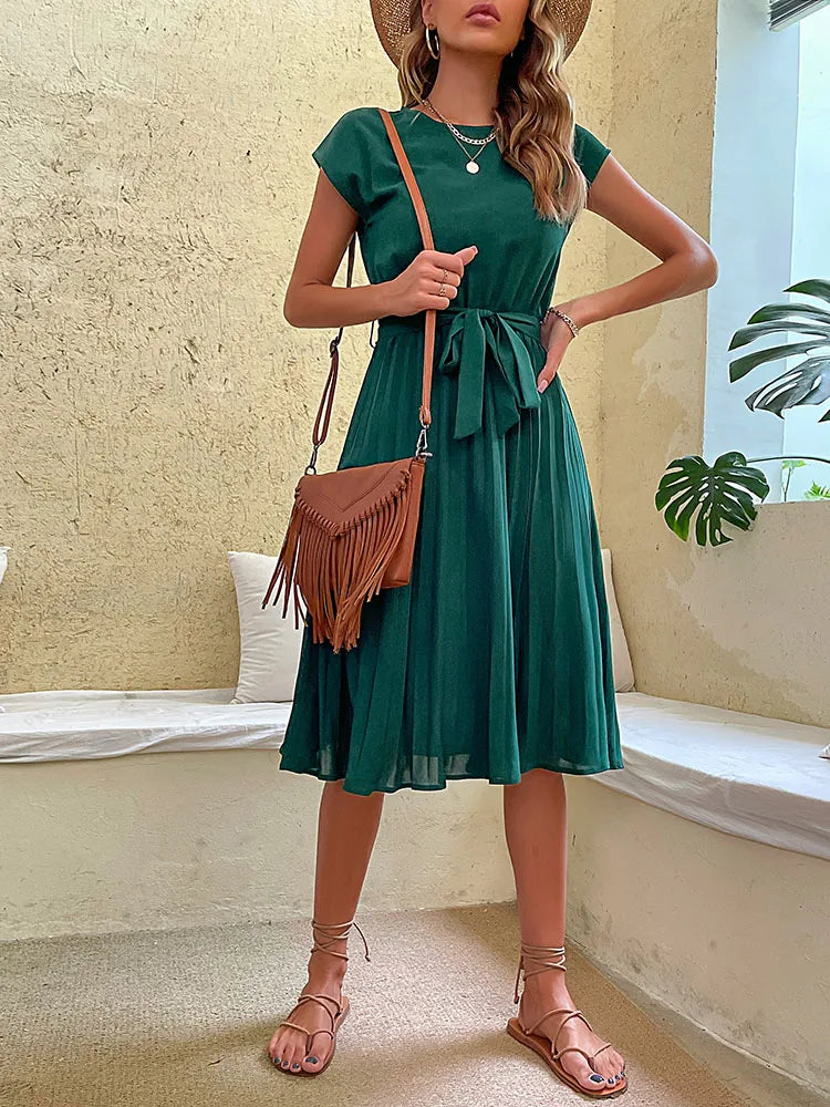 Woman wearing elegant green summer beach dress with tied waist and brown fringe shoulder bag in neutral setting