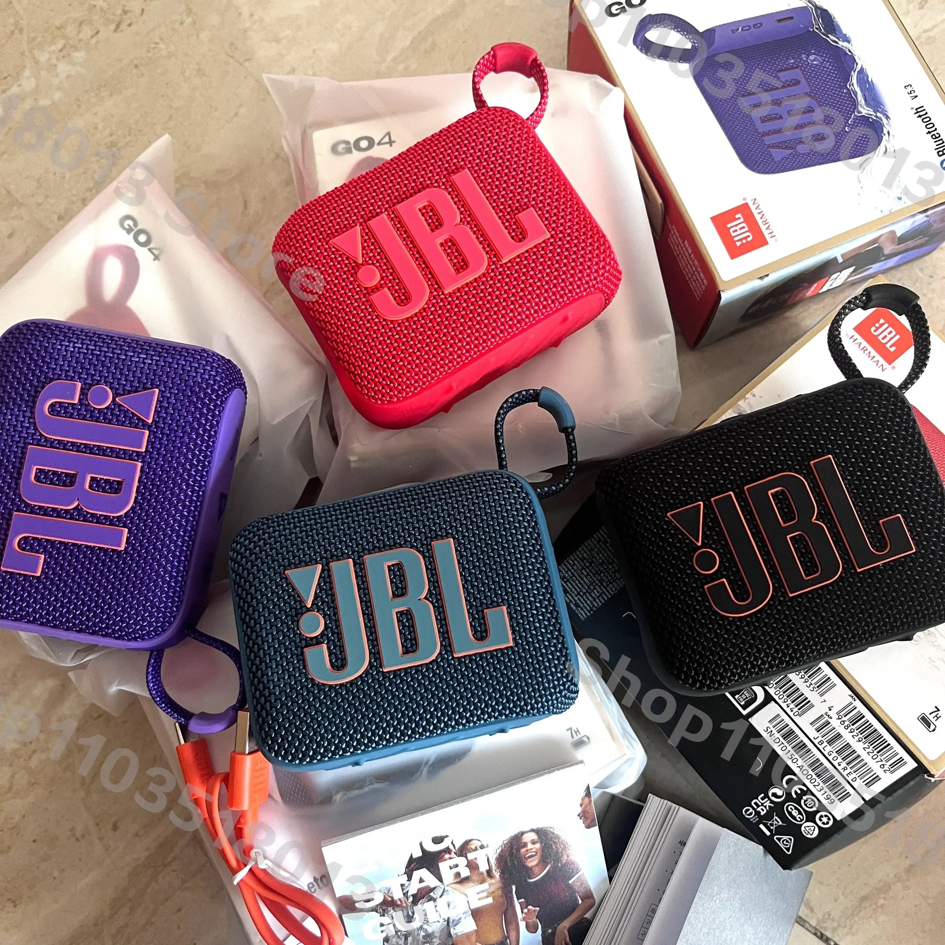 Four JBL GO 4 waterproof portable Bluetooth speakers in purple, red, blue, and black on a table with packaging and accessories