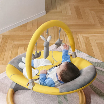 Baby lying in a yellow and gray multifunctional electric rocking chair reaching for hanging soft toys indoors on a wooden floor