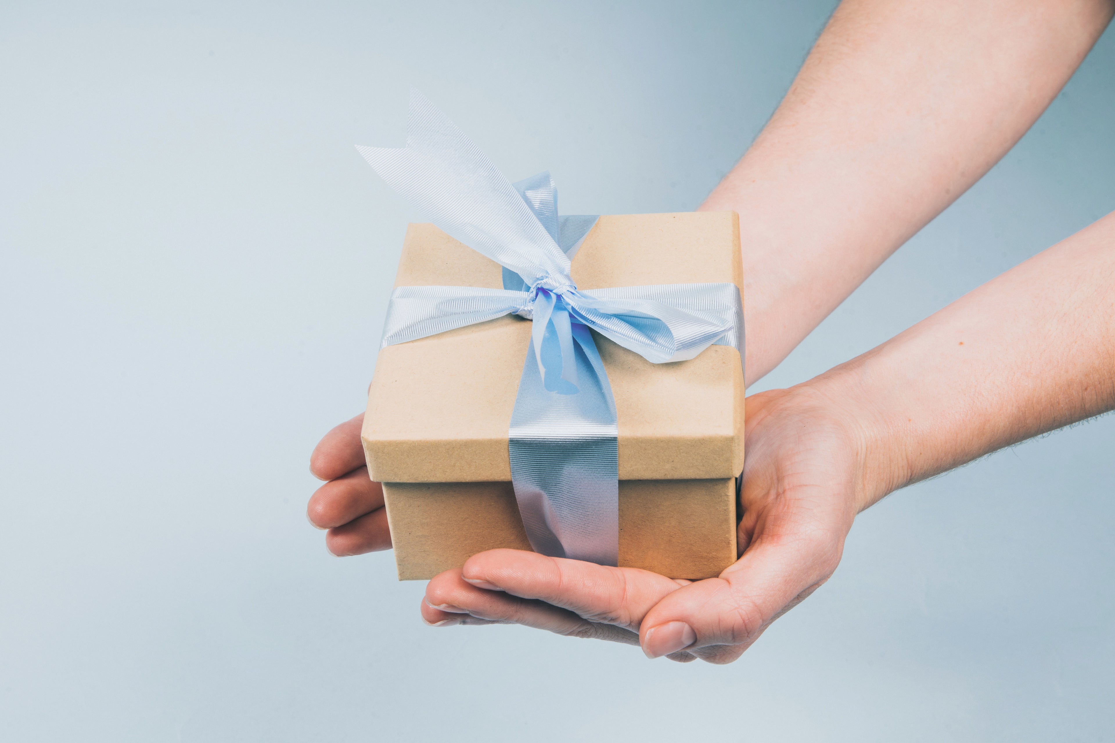 Hands holding a small beige gift box wrapped with a light blue ribbon against a soft blue background