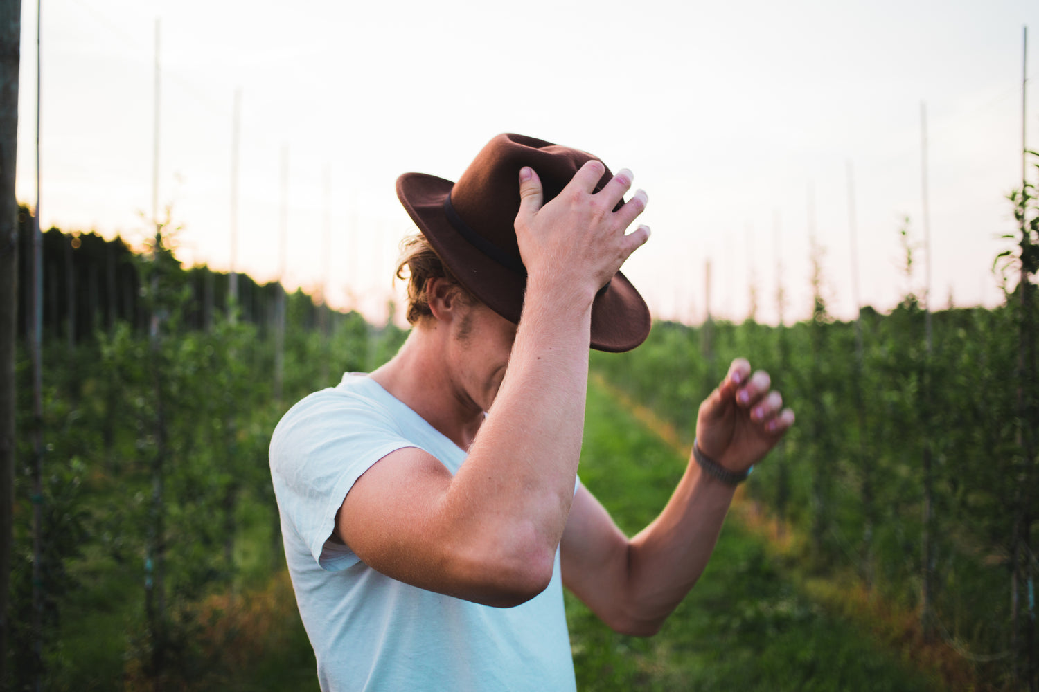 Young man in a white t-shirt adjusting his brown hat standing in a green field during sunset
