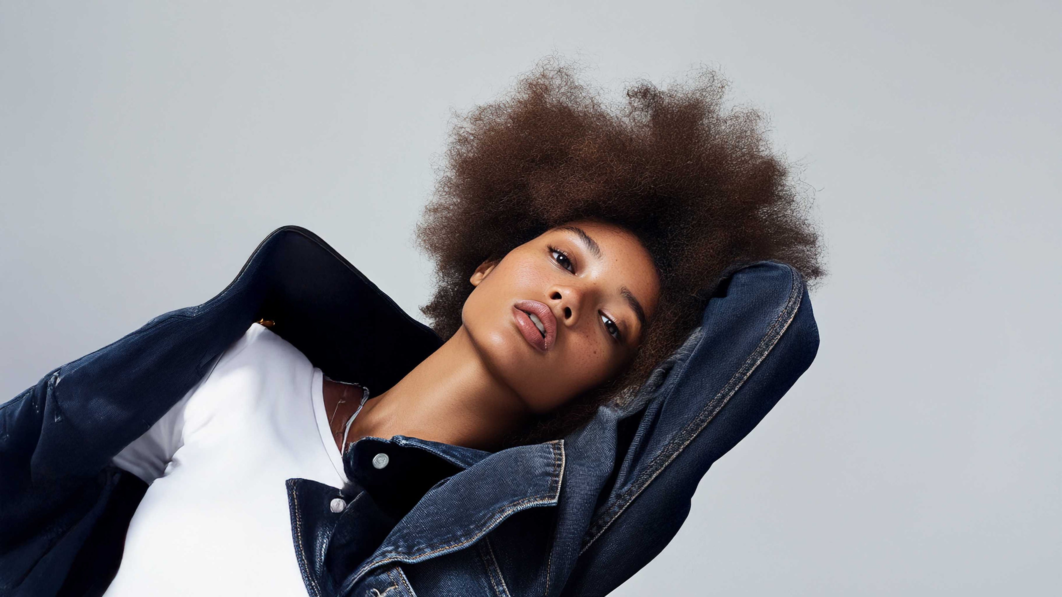 Woman with natural afro hairstyle wearing a white top and dark denim jacket reclining against a light gray background