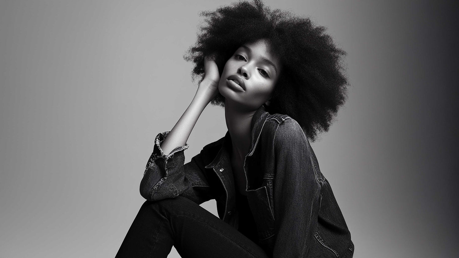 Black and white photo of a woman with natural afro hairstyle wearing a denim jacket and pants seated against a plain background