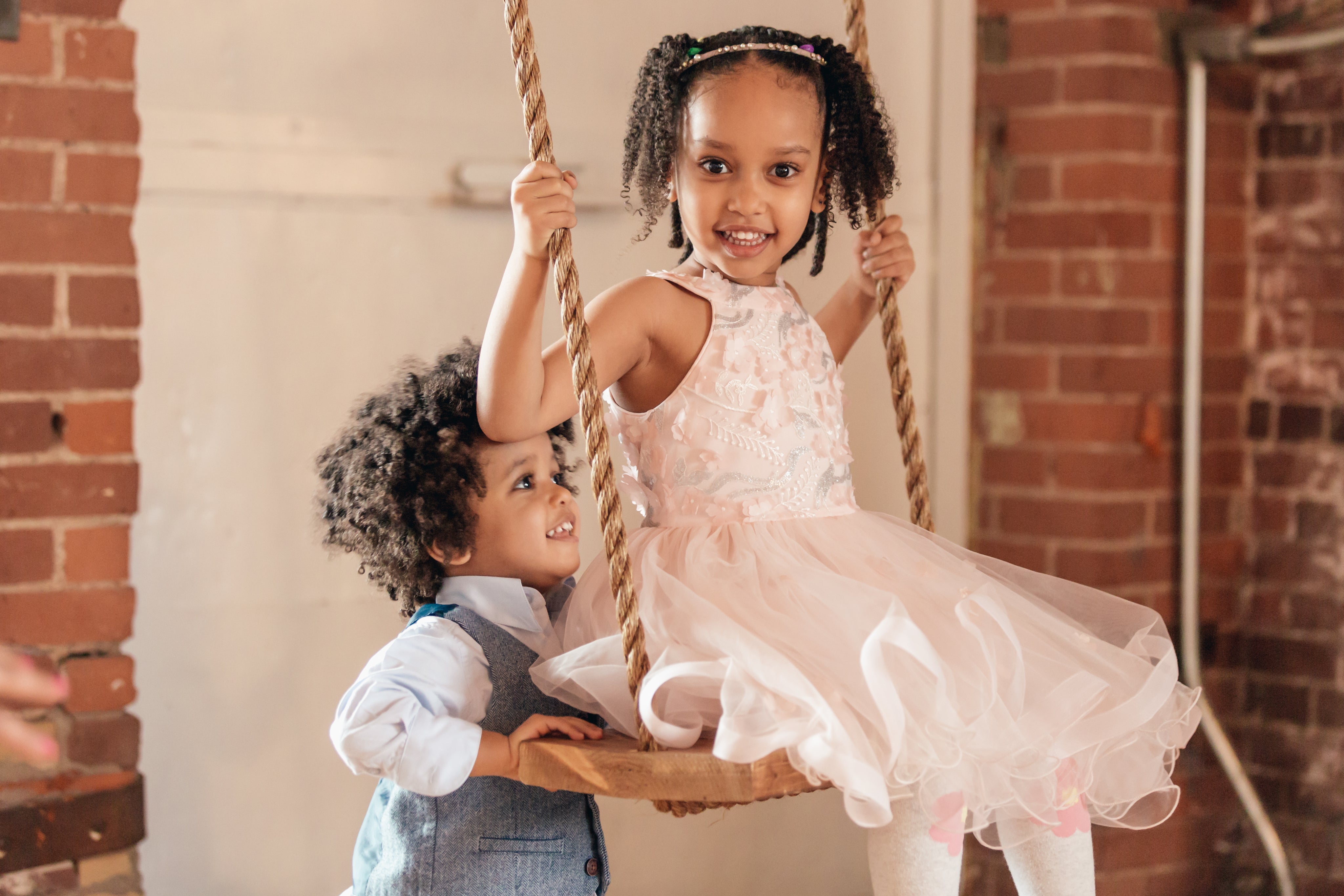 Two young children playing indoors, one sitting on a wooden swing and the other standing beside her smiling