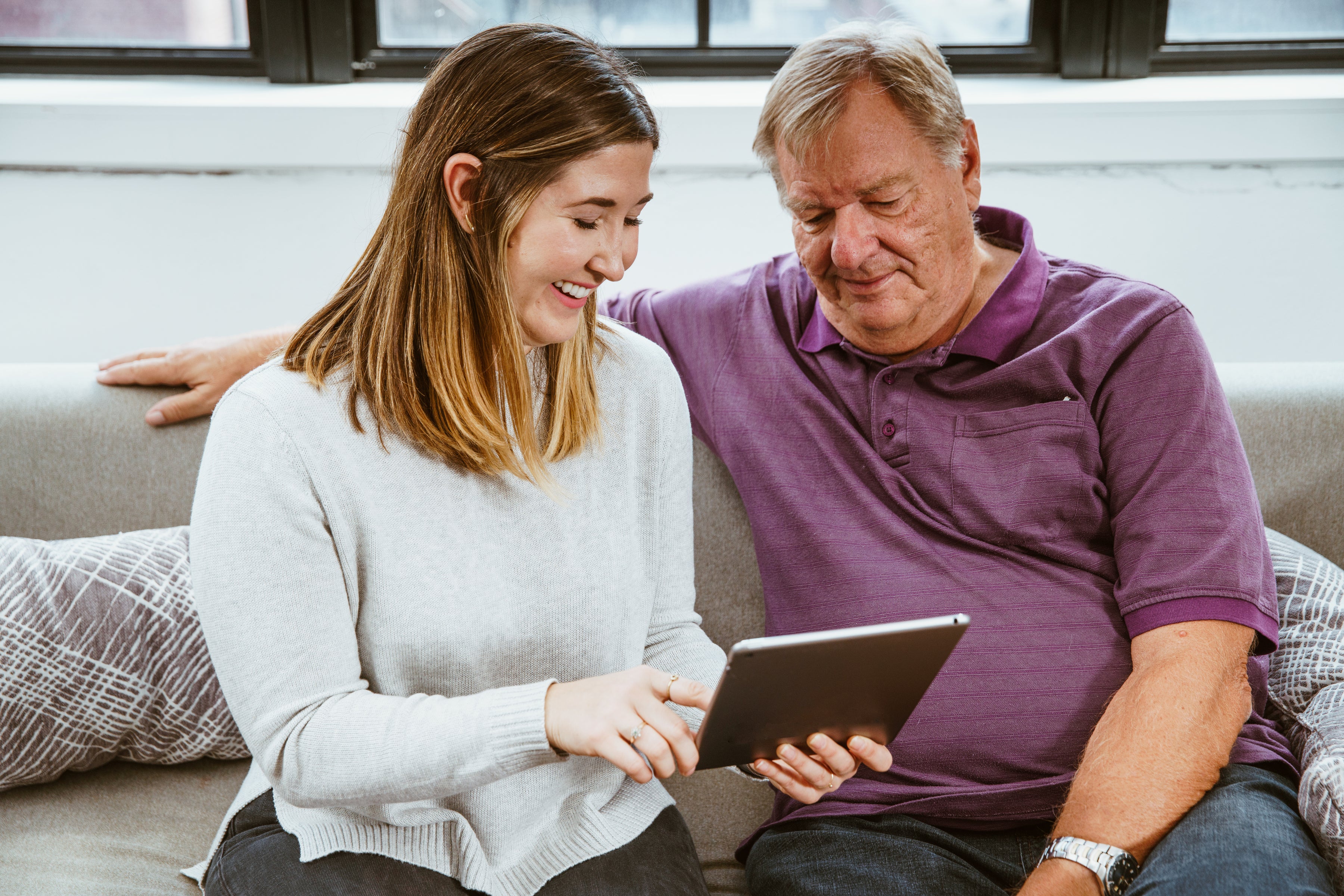 Young woman showing an older man something on a tablet while sitting together on a couch indoors